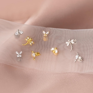 Puede incluir: Una colección de pequeños pendientes decorativos con diseños de insectos y flores. Los pendientes incluyen una abeja, una mariposa, una libélula y un colibrí, con acabados plateados y dorados. Algunos pendientes están adornados con piedras preciosas transparentes.