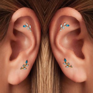 May include: Close-up of two ears, each adorned with a delicate gold earring. The earrings feature a branch design with blue and clear gemstones. The earrings are a stylish accessory.