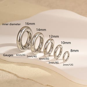 Może przedstawiać: Pięć srebrnych okrągłych pierścieni ułożonych według rozmiaru, oznaczonych średnicami wewnętrznymi 16mm, 14mm, 12mm, 10mm i 8mm. Poniżej podano odpowiadające im grubości: 6mm/2G, 5mm/4G, 4mm/6G, 3mm/8G i 2mm/12G. Każdy pierścień ma gładkie wykończenie.