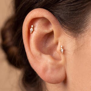 May include: Two gold stud earrings featuring a kite-shaped clear gemstone topped with a small round clear stone. One earring is positioned in the upper cartilage area, while the other is placed in the tragus area of the ear, both set in gold.