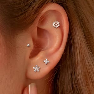 May include: A set of four silver-toned ear piercings featuring a snowflake-shaped stud in the upper cartilage, a small round solitaire stud in the conch, a four-petaled flower stud in the mid-ear, and a five-petaled flower stud in the earlobe.