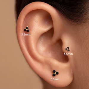 Può includere: Tre orecchini a lobo color oro, ciascuno caratterizzato da un gruppo di tre pietre nere disposte a triangolo. Gli orecchini sono etichettati con le misure 5,5 mm, 4,5 mm e 6,5 mm, che indicano le dimensioni specifiche di ogni pezzo.