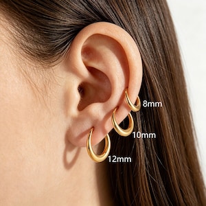 Puede incluir: Tres pendientes de aro dorados en una sola oreja, con tamaños variables etiquetados como 8mm, 10mm y 12mm. Los aros de metal liso y pulido están dispuestos en una fila vertical a lo largo del lóbulo y el cartílago, mostrando un diseño simple.
