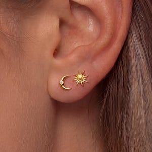 Puede incluir: Un par de pendientes de botón en tono dorado que presentan una forma de media luna con una pequeña piedra clara incrustada y una forma de sol con rayos radiantes. Los pendientes están colocados en el lóbulo de la oreja con acabado metálico.
