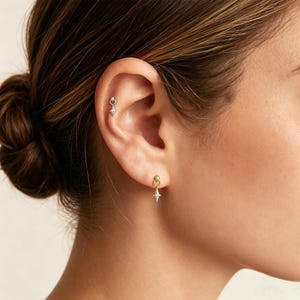 May include: A silver-toned star-shaped charm earring with a small ball stud, worn in the earlobe, and a matching silver-toned star-shaped charm earring with a small ball stud, worn in the upper cartilage area of the ear.