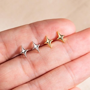 May include: Two pairs of star-shaped stud earrings are shown. One pair is silver, and the other is gold. The earrings are small and have four points. The earrings are displayed on a person's palm.