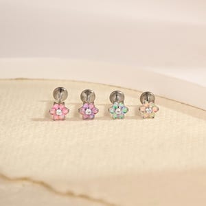 Puede incluir: Cuatro pendientes de botón con parte posterior plana, cada uno con un diseño en forma de flor. Los pétalos están compuestos por pequeñas piedras cabujón redondas e iridiscentes en tonos rosa, morado, azul y blanco, con una piedra clara en el centro.