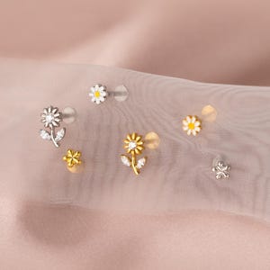 May include: A collection of daisy-shaped stud earrings in silver and gold tones. Some earrings feature clear crystals and white enamel detailing. The earrings are displayed on a soft, pink fabric background.
