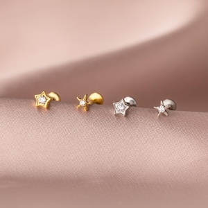 Könnte beinhalten: Vier sternförmige Ohrstecker in Gold- und Silbertönen, jeweils mit einem kleinen, klaren Edelstein in der Mitte. Die Ohrringe werden auf einer rosa, strukturierten Oberfläche präsentiert, die ihr zartes Design und ihren Glanz hervorhebt.
