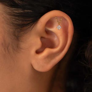 May include: A gold ear piercing with a four-petal flower design set with clear stones. The earring hangs from a fine chain, adding a touch of elegance. The jewelry is shown on an ear.