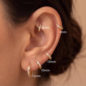 May include: A set of gold hoop earrings with embedded clear stones, displayed in various ear piercings. The hoops are labeled with sizes 6mm, 8mm, 10mm, and 12mm, showing a double-band design where one band is plain gold and the other features stones.
