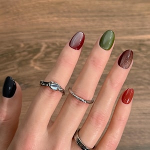 May include: Three silver-toned rings worn on fingers. One ring features a central light blue stone, another is a plain thin band, and the third is a wider band with engraved text. The fingernails are painted in various colors: black, dark red, olive green, brown, and brick red.