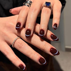 Könnte beinhalten: Ovale Nägel, die in einem tiefen Burgunderrot lackiert sind, mit einem dünnen, hellblauen Halbmond-Design in der Nähe der Nagelhaut. Die Hände zeigen auch mehrere Ringe, darunter ein Silberband, ein diamantbesetzter Ring und ein Ring mit einem großen blauen Edelstein.