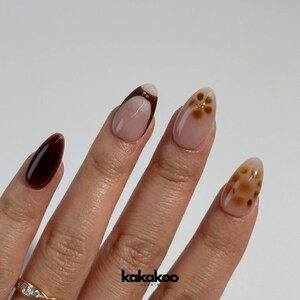 May include: Close-up of a hand with almond-shaped nails painted in a brown and white ombre design. One nail features a brown and gold floral design, another has a brown and gold leopard print design, and another is solid brown.