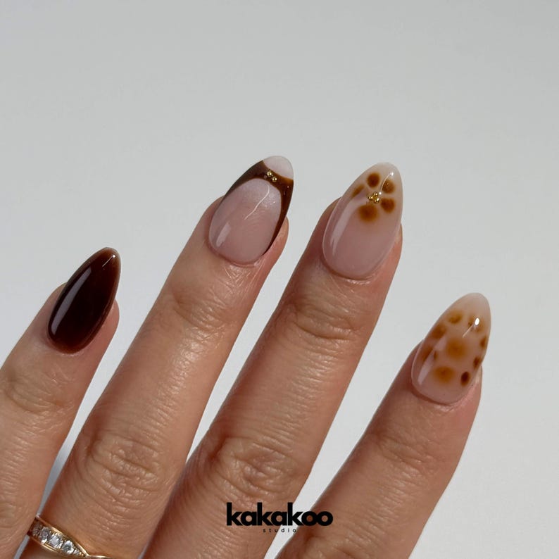 May include: Close-up of a hand with almond-shaped nails painted in a brown and white ombre design. One nail features a brown and gold floral design, another has a brown and gold leopard print design, and another is solid brown.