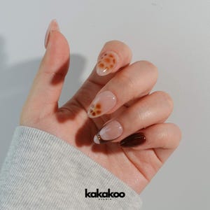 May include: Close-up of a hand with five nails painted with different designs. The nails are painted with a light brown, white, and brown color. The nails have a floral design and a brown and white spotted design.