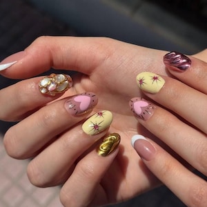 Op de afbeelding: Een hand met verschillende nail art-ontwerpen. De nagels hebben een gouden ring, een French manicure en nagels beschilderd met harten, sterren en metallic afwerkingen in roze, geel en goud.