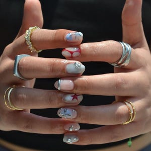 May include: Close-up of manicured nails with various designs. Some nails feature a red star, silver embellishments, and a bikini design. The nails are adorned with a variety of rings in gold and silver.