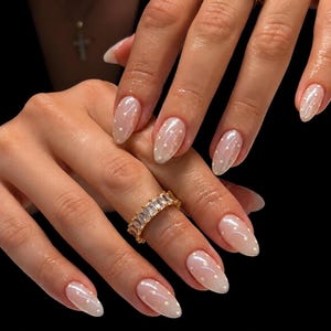 Puede incluir: Primer plano de uñas manicuradas con un acabado claro nacarado y lunares blancos. Un anillo dorado con piedras claras rectangulares adorna un dedo. Las uñas son almendradas y el aspecto general es elegante.