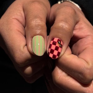 May include: Close-up of two manicured fingernails. One nail is light green with vertical pink stripes. The other nail is a pink and burgundy checkerboard pattern, adorned with two small gold beads. The nails are on a hand.