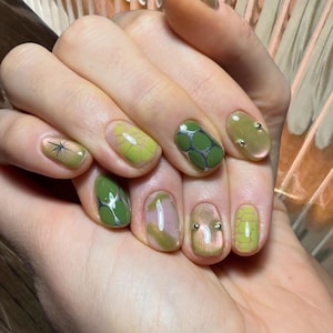 Puede incluir: Una mano con una manicura que presenta varios dise&ntilde;os de u&ntilde;as verdes. Algunas u&ntilde;as tienen un patr&oacute;n floral, otras un dise&ntilde;o geom&eacute;trico, y algunas est&aacute;n adornadas con peque&ntilde;os tachuelas plateadas. Las u&ntilde;as est&aacute;n pintadas en tonos verdes y un rosa claro.