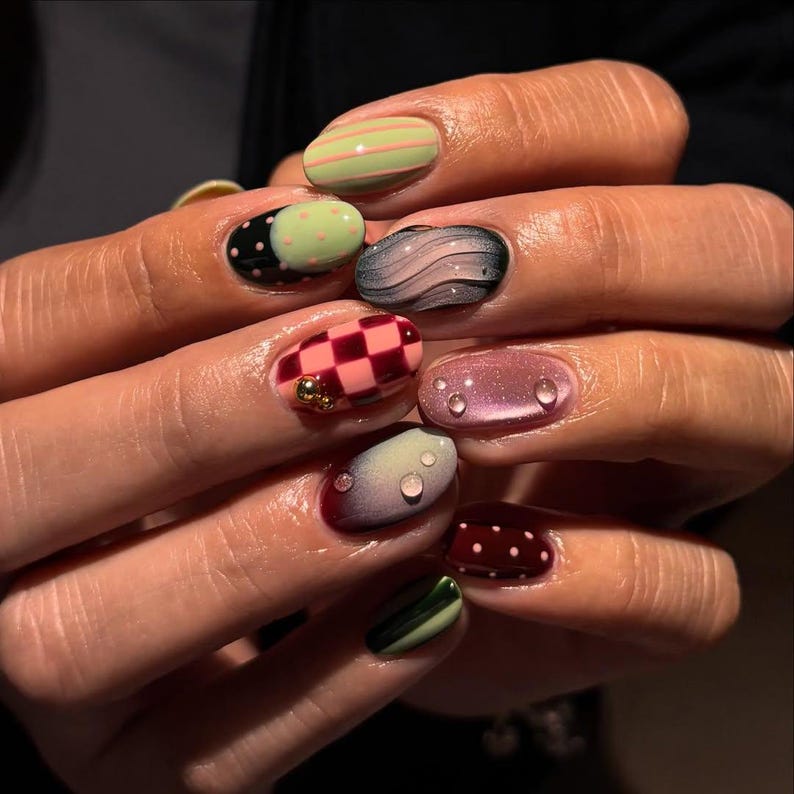 May include: A close-up of a hand with a variety of manicured nails. The nails feature designs such as stripes, polka dots, checkerboard patterns, and water droplet effects. Colors include green, pink, black, and burgundy.