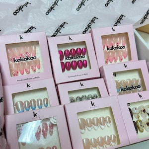 May include: A selection of pink boxes displaying press-on nail sets. The nail designs include glitter, floral patterns, and solid colours. The brand name "kakakoo studio" is printed on the boxes.