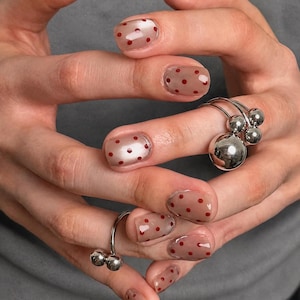 May include: Nails featuring a sheer nude base coat with small, evenly spaced red polka dots. Two silver rings are worn, each characterised by a cluster of polished spherical beads of varying sizes attached to a thin, open-ended metal band.