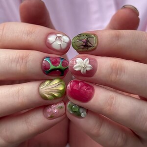 May include: A set of fingernails featuring intricate 3D nail art designs. The nails display a variety of textures and motifs, including raised white flowers, metallic green shell-like shapes, translucent pink layers, and abstract green and red patterns.