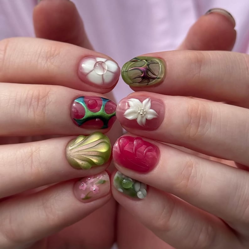 May include: A set of fingernails featuring intricate 3D nail art designs. The nails display a variety of textures and motifs, including raised white flowers, metallic green shell-like shapes, translucent pink layers, and abstract green and red patterns.