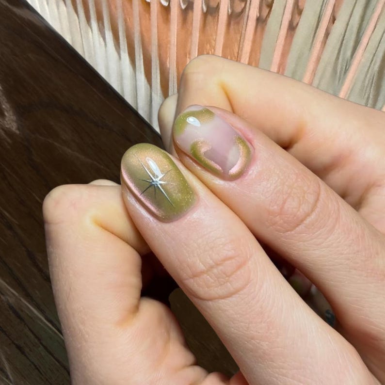 Puede incluir: Primer plano de u&ntilde;as manicuradas con un dise&ntilde;o de estrella y esmalte dorado y verde oliva. Una u&ntilde;a presenta una estrella plateada, mientras que otras tienen remolinos dorados sobre una base nude. Las u&ntilde;as est&aacute;n bien cuidadas y son elegantes.