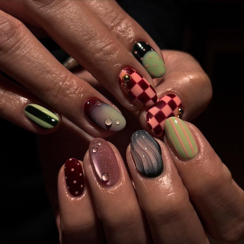 May include: A close-up of a hand with a variety of manicured nails. The nails feature designs such as checkered patterns, stripes, polka dots, and gradient effects. Colors include red, pink, green, black, and blue. Some nails have small, decorative embellishments.