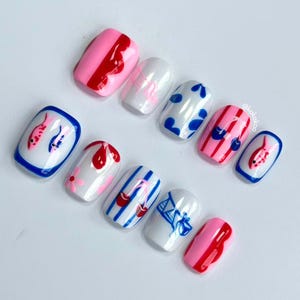 Op de afbeelding: Een set van tien kunstnagels met diverse ontwerpen. De nagels hebben een mix van roze, witte en blauwe kleuren. Sommige nagels hebben rode en blauwe patronen, waaronder vissen, kersen en stippen. De nagels zijn gerangschikt op een wit oppervlak.