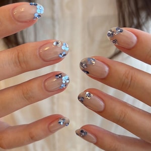 May include: A manicure featuring a clear, glossy base coat on oval-shaped nails. Each nail is decorated with small, delicate blue and silver floral-shaped sequins or charms placed near the tips, creating a subtle and reflective accent.