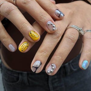 Op de afbeelding: Gemanicureerde vingernagels met diverse ontwerpen, waaronder felgele nagellak met zwarte stippen, transparante lak met zwarte lijnaccenten, transparante lak met kleine zilveren metalen bolletjes en één lichtblauwe iriserende nagel.