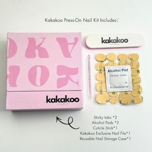 May include: A pink Kakakoo press-on nail kit with a box, nail file, alcohol pads, cuticle stick, and sticky tabs. The box has the brand name and logo in black and pink. The kit includes a reusable nail storage case.