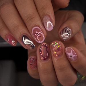 May include: A hand with a manicure featuring various nail art designs. The nails showcase a mix of pink, silver, and clear gel, with star, heart, and abstract metallic accents. Some nails have glitter or a glossy finish.