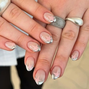 May include: A close-up of manicured nails with a clear base, silver tips, and pink star designs. The nails are oval-shaped and feature a modern, minimalist style. The image also shows two silver rings on the fingers.