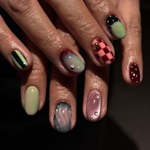May include: A close-up of a hand with a variety of manicured nails. The nails feature different designs, including stripes, polka dots, and checkered patterns. Colors include green, pink, red, and black. Some nails have water droplet accents.