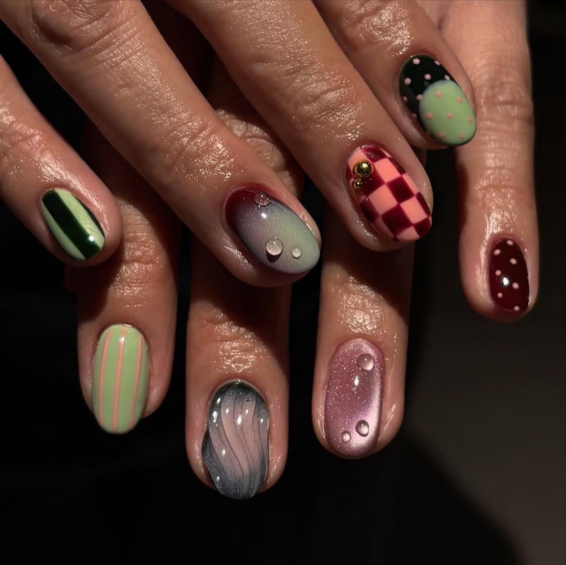 May include: A close-up of a hand with a variety of manicured nails. The nails feature different designs, including stripes, polka dots, and checkered patterns. Colors include green, pink, red, and black. Some nails have water droplet accents.