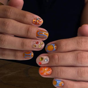 Peut inclure: Gros plan sur une manucure présentant divers motifs de nail art. Les ongles présentent un mélange de vernis orange, bleu et transparent avec des volutes abstraites, et sont ornés de petits bijoux et paillettes colorés. L'esthétique générale est moderne et artistique.