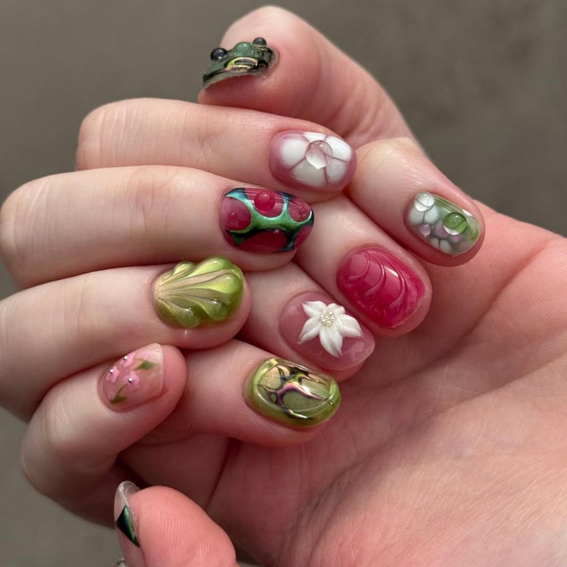 May include: A set of fingernails featuring diverse 3D nail art designs including a small green frog figure, white floral accents, pink and green abstract patterns, textured shell-like shapes, and small pink blossoms with tiny silver beads.