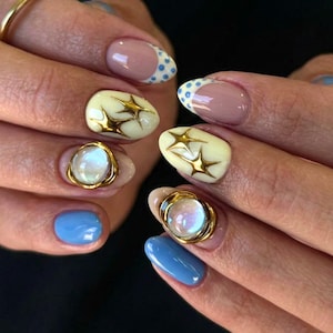 Op de afbeelding: Gemanicureerde vingernagels met een mix van ontwerpen, waaronder lichtblauwe lak, French tips met blauwe stippen, crèmekleurige nagels met gouden steraccenten en nagels versierd met verhoogde, iriserende ronde cabochons in gouden zettingen.