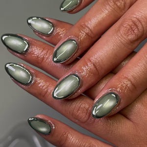 May include: Close-up of a hand with oval-shaped, light green nails. Each nail has a silver metallic border, creating a unique, modern manicure design. The nails have a glossy finish, reflecting light.