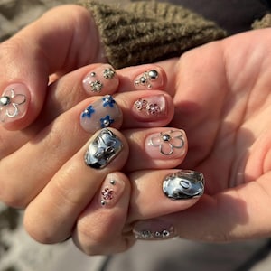 May include: A hand with a manicure featuring a variety of nail art. Some nails have silver flower designs, others have blue flower patterns, and some have silver metallic accents. The nails are painted in a range of colors, including clear, pink, and silver.