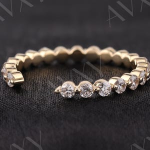 May include: A gold-colored ring with a row of clear, round gemstones. The ring has a delicate design, with the gemstones set closely together. The ring is displayed on a dark, textured surface, highlighting its sparkle and elegance.