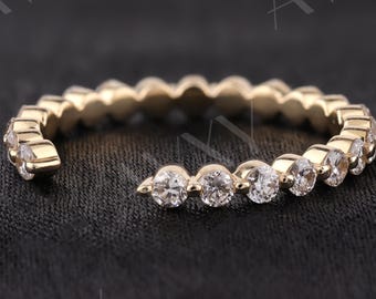 May include: A gold-colored ring with a row of clear, round gemstones. The ring has a delicate design, with the gemstones set closely together. The ring is displayed on a dark, textured surface, highlighting its sparkle and elegance.