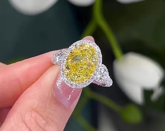 May include: A dazzling ring featuring a large, oval-cut yellow gemstone, surrounded by a halo of smaller, clear stones. The ring's band is also adorned with clear stones, creating a sparkling effect. The ring is set in a silver or white metal.