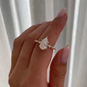 May include: A pear-shaped diamond engagement ring with a gold band and smaller diamonds set around the main stone.