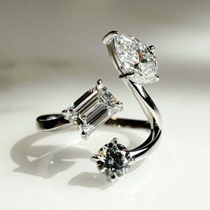 May include: A silver ring with three diamonds. The ring has a curved band and the diamonds are set in different shapes, including a pear, an emerald cut, and a round brilliant.
